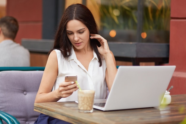 Tiro horizontal de freelancer feminino ocupado mantém telefone celular, mensagens de texto on-line, usa computador portátil, bebe milk-shake, posa em restaurante ao ar livre, vestido com roupas brancas