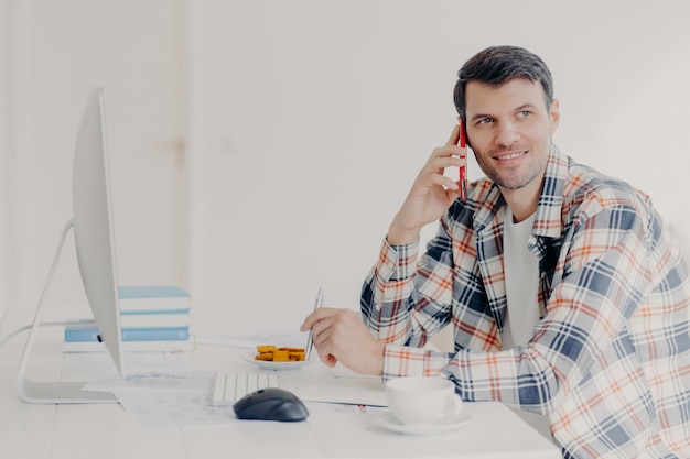 Tiro horizontal de freelancer masculino ocupado tem conversa telefônica, vestido com camisa quadriculada, anota informações, senta-se na área de trabalho com computador, resolve problema financeiro, posa no local de trabalho