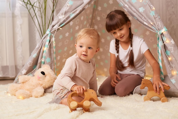 Tiro horizontal de irmão otimista positivo aproveitando o tempo em casa menina com tranças brincando com brinquedos de madeira na tenda junto com sua irmã criança