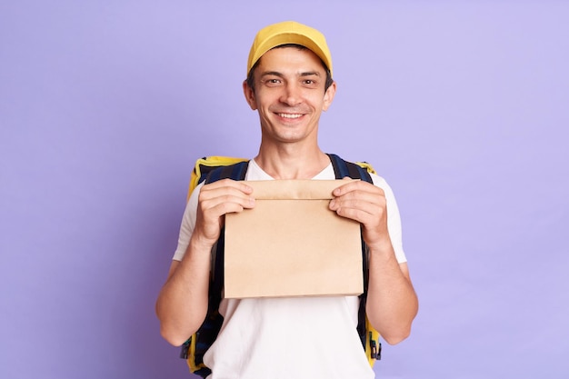 Tiro horizontal de jovem adulto bonito entregador caucasiano segurando saco de papel com comida para viagem muito feliz olhando para a câmera com expressão positiva