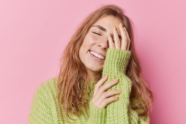 Tiro horizontal de jovem alegre feliz inclina a cabeça cobre o rosto com a mão sorri amplamente mantém os olhos fechados ri de algo positivo usa suéter de inverno isolado sobre fundo rosa.