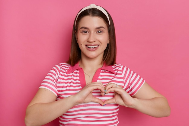 Foto tiro horizontal de jovem romântica feliz vestindo camiseta listrada e faixa de cabelo mostrando gesto de coração com as mãos expressando amor e gentil posando isolado sobre fundo rosa