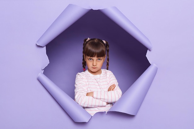 Tiro horizontal de menina adorável triste ofendida com tranças olhando através do avanço do fundo roxo em pé com as mãos postas expressando tristeza