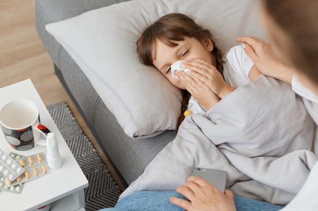 Tiro horizontal de menina doente com nariz escorrendo e segurando a criança de tecido deitada na cama debaixo do cobertor e sentindo-se mal tendo sintomas de alta temperatura e gripe