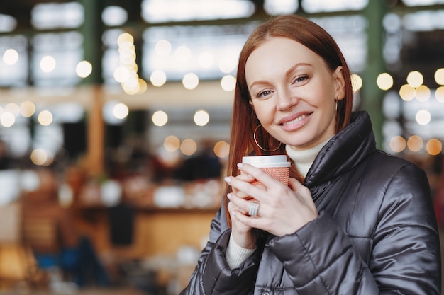 Tiro horizontal de mulher adorável com sorriso, segura o copo de papel da bebida, goza de café aromático