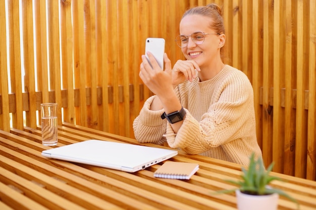 Tiro horizontal de mulher adulta jovem e bonita com penteado de coque vestindo suéter bege segurando o telefone inteligente nas mãos tendo chamada de vídeo ou fazendo selfie