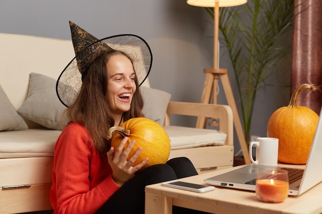 Foto tiro horizontal de mulher animada e feliz vestindo suéter e chapéu de bruxa segurando abóbora laranja nas mãos e conversando por videochamada com seus amigos rindo alegremente durante o feriado de halloween