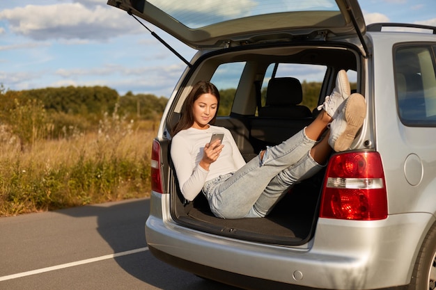 Tiro horizontal de mulher bonita de cabelos escuros sentado no porta-malas aberto de um carro e usando o celular para verificar as redes sociais enquanto descansa da condução