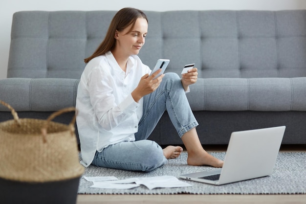 Tiro horizontal de mulher caucasiana cativante vestindo camisa branca e jeans sentado no chão perto do sofá usando laptop e telefone segurando o cartão de crédito olhando para a tela