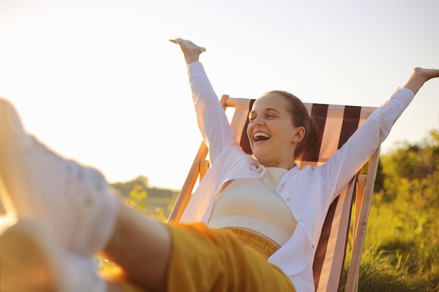 Tiro horizontal de mulher caucasiana de cabelos escuros extremamente feliz vestindo camisa branca sentada na cadeira dobrável pela água levantou os braços olhando belas vistas gritando de felicidade