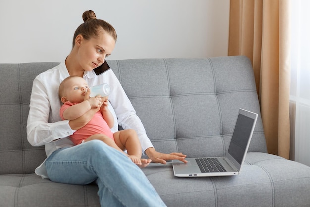 Tiro horizontal de mulher com penteado de coque vestindo camisa branca e jeans sentado na tosse segurando a filha nas mãos falando via celular e digitando no laptop