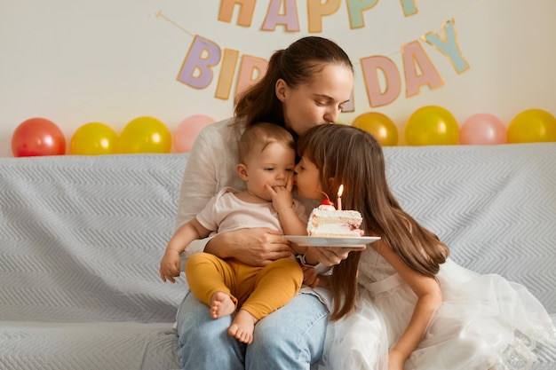 Tiro horizontal de mulher com suas filhas sentadas na tosse com bolo de aniversário comemorando férias posando com balões e inscrição festiva no fundo mamãe beijando seus filhos