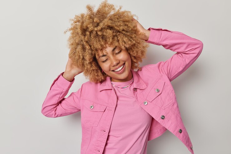 Foto tiro horizontal de mulher de cabelo encaracolado alegre despreocupada mantém as mãos atrás da cabeça mantém os olhos fechados sorrisos amplamente vestido com elegante jaqueta rosa isolada sobre fundo branco tem sonhos agradáveis