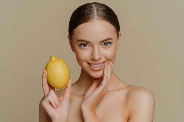 Tiro horizontal de mulher europeia bastante saudável usa limão para fazer máscara facial natural cuida de seu corpo e pele fica em topless interior isolado sobre fundo marrom Conceito de beleza