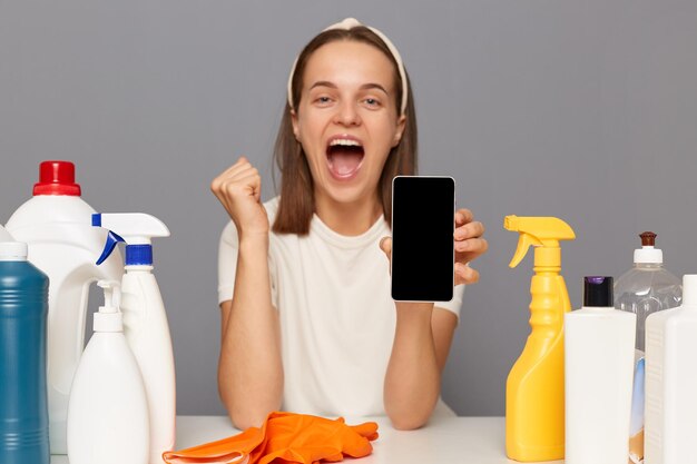 Tiro horizontal de mulher extremamente feliz usando luvas de proteção senta-se à mesa com muitos detergentes posando isolado sobre fundo cinza mostrando telefone celular com tela vazia