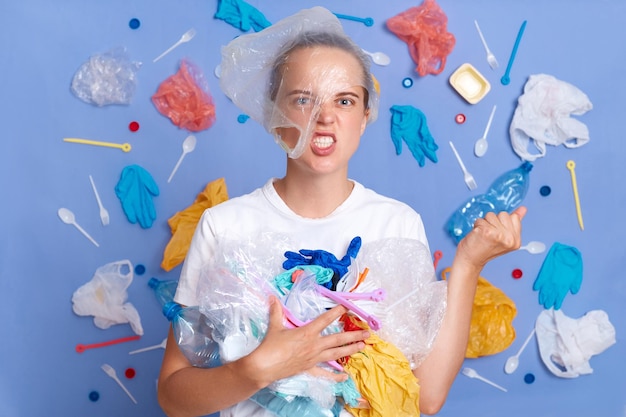 Tiro horizontal de mulher irritada com pacote de plástico na cabeça mostra punho exigindo de pessoas ecologicamente amigáveis tem um olhar estrito em pé contra a parede azul com muito lixo