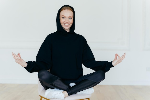 Tiro horizontal de mulher otimista pratica ioga medita na cadeira senta-se em pose de lótus usa moletom preto tênis branco tem expressão feliz tenta relaxar após um árduo dia de trabalho