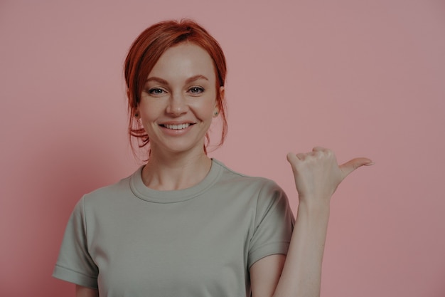 Tiro horizontal de mulher ruiva alegre com expressão facial feliz, indicando com o dedo no espaço em branco da cópia, mulher apontando com o polegar enquanto anuncia o produto. Conceito de propaganda