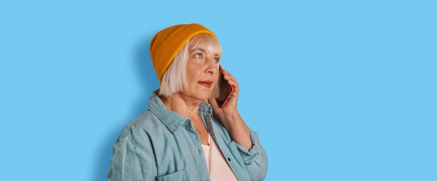 Tiro horizontal de mulher sênior moderna alegre usa telefone falando em um telefone celular tem humor feliz sobre fundo de cor azul