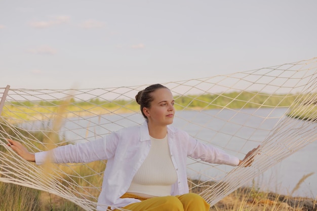 Tiro horizontal de mulher vestindo camisa branca sentada na rede na margem do rio olhando para longe com expressão sonhadora posando com céu e água no fundo