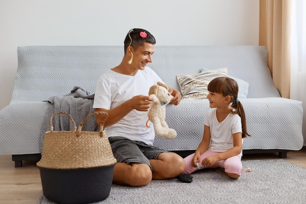 Tiro horizontal de pai adulto jovem satisfeito, passando um tempo com sua filha enquanto está sentado no chão perto do sofá na sala de estar, pai segurando um brinquedo macio de cachorro, família se divertindo e brincando juntos.