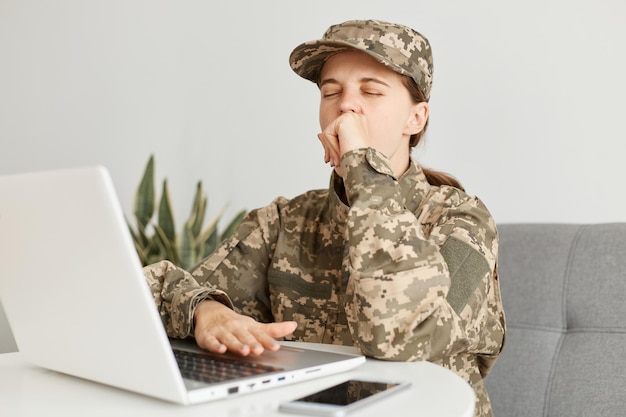 Tiro horizontal de soldado mulher com sono usando chapéu e uniforme de camuflagem, posando em casa, trabalhando online no computador portátil, sente-se cansado, bocejando, mantém os olhos fechados.