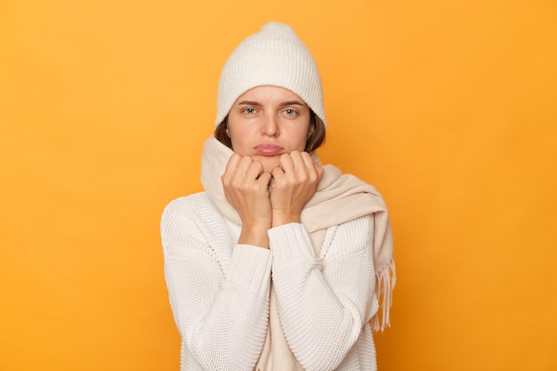 Tiro horizontal de triste mulher caucasiana chateada vestindo boné quente e cachecol posando isolado sobre fundo amarelo em pé com lábios beicinho de mau humor