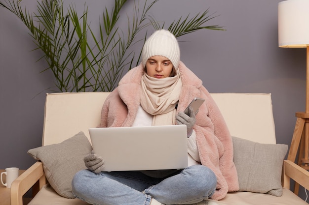 Tiro horizontal de triste mulher caucasiana infeliz vestindo boné luvas cachecol e casaco sentado no sofá com laptop e telefone inteligente nas mãos sendo ocupado posando na sala fria