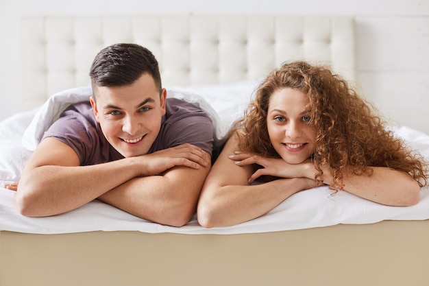 Tiro horizontal do casal satisfeito satisfeito da família deitar na cama confortável, estar de bom humor após a noite inesquecível. Linda mulher encaracolada encontra-se perto de seu namorado. Conceito de relacionamento.