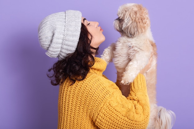 Tiro horizontal do estúdio da moça alegre atrativa que está isolada sobre o lilás, mantendo seu cão maltês nas mãos, beijando-o, amando o animal de estimação muito.