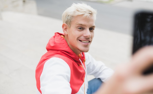 Tiro horizontal do homem elegante jovem loiro hipster sorrindo e vestindo capuz com zíper na moda tirar autoretrato com telefone inteligente ao ar livre no parque pessoas estilo de vida e conceito de tecnologia