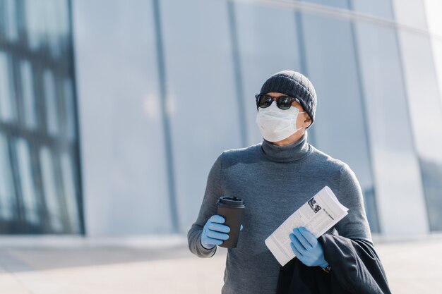 Tiro horizontal do homem em perigo de pegar coronavírus em local público, protege-se usando máscara médica e luvas de borracha, bebe café para viagem, olha para algum lugar através de óculos de sol
