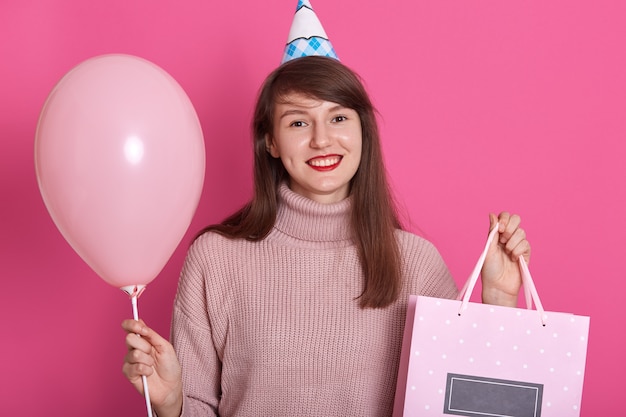 Tiro horizontal de encantadora mujer sonriente viste suéter rosa y cono