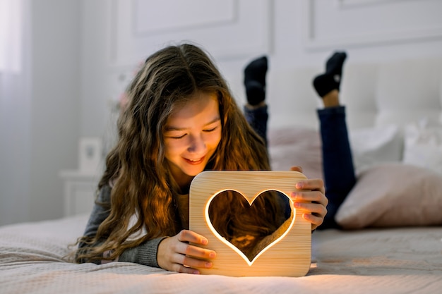 Tiro horizontal interior de linda menina da escola infantil, deitada na cama em casa e se divertindo com a elegante lâmpada noturna de madeira feita à mão com imagem de coração.
