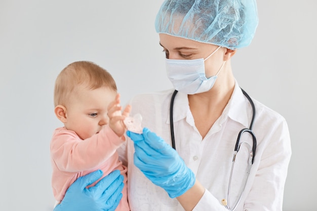 Tiro horizontal del médico de familia sonriente mujer positiva con uniforme médico y mascarilla quirúrgica sosteniendo al bebé en las manos, informa a los padres sobre la salud del niño.