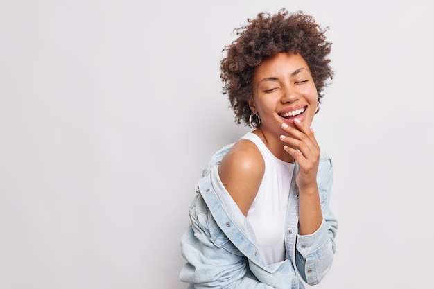 Tiro horizontal de mujer alegre y llena de alegría con cabello rizado sonríe ampliamente se siente muy feliz y optimista expresa auténticas emociones positivas mantiene los ojos cerrados usa chaqueta de mezclilla camiseta blanca