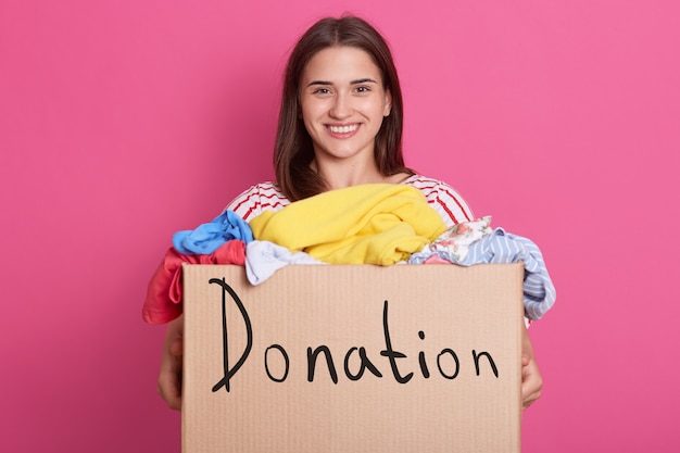 Foto tiro horizontal de la situación femenina voluntaria aislada sobre rosa, caja que se sostiene con la donación de la inscripción, ropa para el hogar del `s de los niños. mujer morena sonriente que hace caridad.