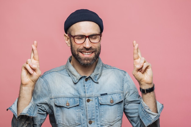 Tiro indoor de bonito homem com barba mantém os dedos cruzados, olhos fechados, tem expressão feliz