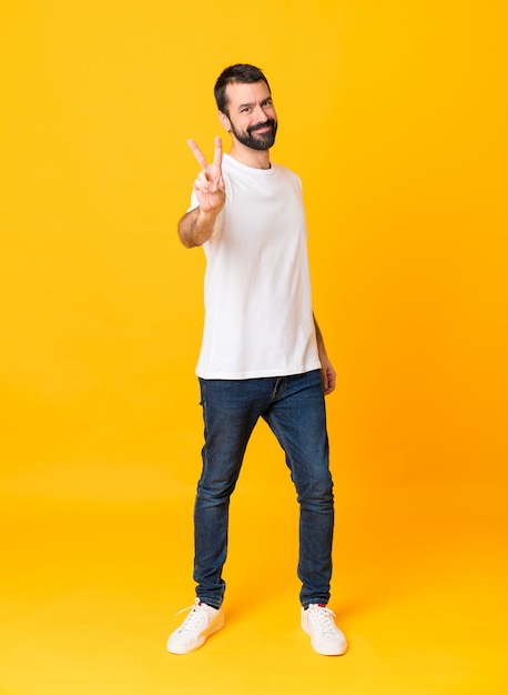 Tiro integral del hombre con barba sobre amarillo aislado sonriendo y mostrando el signo de la victoria