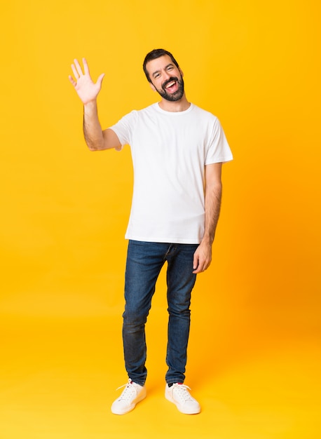 Tiro integral del hombre con barba sobre fondo amarillo aislado saludando con la mano con expresión feliz