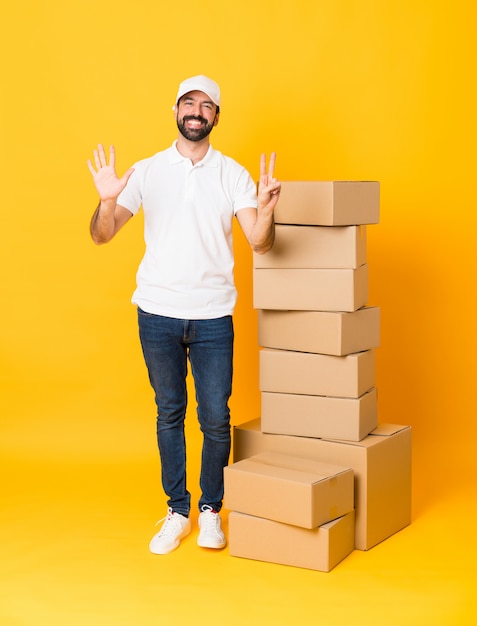 Tiro integral del repartidor entre cajas sobre amarillo aislado contando siete con los dedos