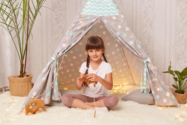 Tiro interior de encantadora garotinha sorridente com tranças vestindo camiseta branca posando em wigwam e brincando com brinquedo ecológico de madeira olhando para a câmera e expressando emoções positivas