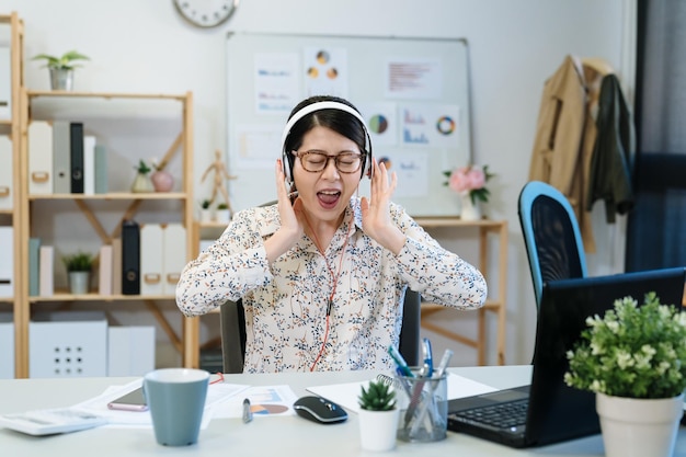 Tiro interior de feliz empresária engraçada cantar e ouvir música favorita através de fones de ouvido estéreo. senhora trabalhadora finge ser cantora de verdade usa fones de ouvido no local de trabalho. funcionária de escritório despreocupada.