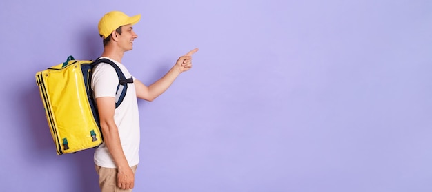 Tiro interior de homem de correio sorridente em camiseta boné e mochila de saco de comida térmica apontando para a área de propaganda expressando emoções positivas isoladas no fundo roxo