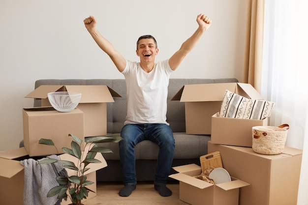 Tiro interior de homem moreno extremamente feliz vestindo camiseta branca e jeans sentado na tosse cercado com uma pilha de pacotes, braços levantados, comemorando a mudança para um novo apartamento ou casa.