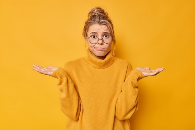 Tiro interior de jovem hesitante e sem noção espalha palmas das mãos não pode tomar decisão encolhe os ombros vestido com jumper casual isolado sobre fundo amarelo modelo feminino questionado se sente incerto