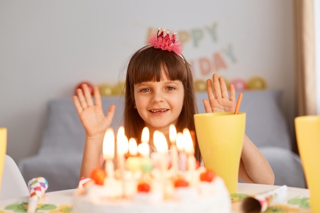 Tiro interior de menina bonitinha otimista feliz com flor no cabelo, sentado à mesa com bolo de aniversário, olhando para a câmera com sorriso feliz, acenando com as mãos