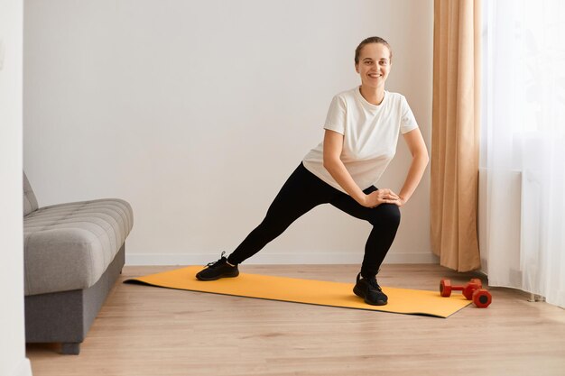 Tiro interior de mulher atlética atraente vestindo camiseta branca e leggins pretas fazendo exercícios esportivos em casa esticando as pernas fazendo exercícios de ioga olhando sorrindo para a câmera