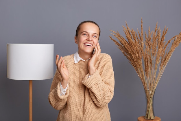 Tiro interior de mulher caucasiana atraente vestindo jumper bege posando em quarto elegante tendo uma conversa agradável em pé e falando telefone com sorriso e rindo de bom humor