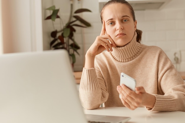 Tiro interior de mulher pensativa triste vestindo suéter bege posando na cozinha segurando o telefone inteligente e olhando para a câmera com expressão facial pensativa